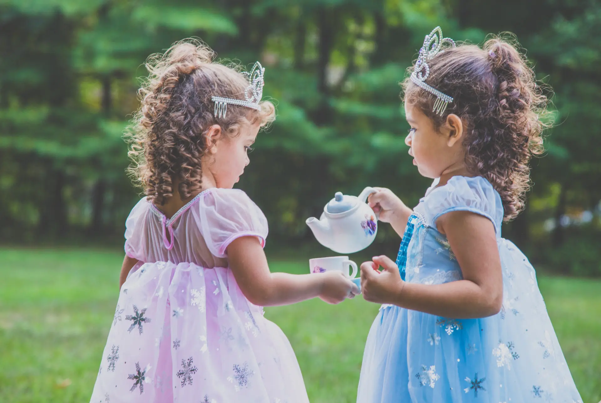 Two little girls in princess dresses and tiaras pouring a tea party in the grass
