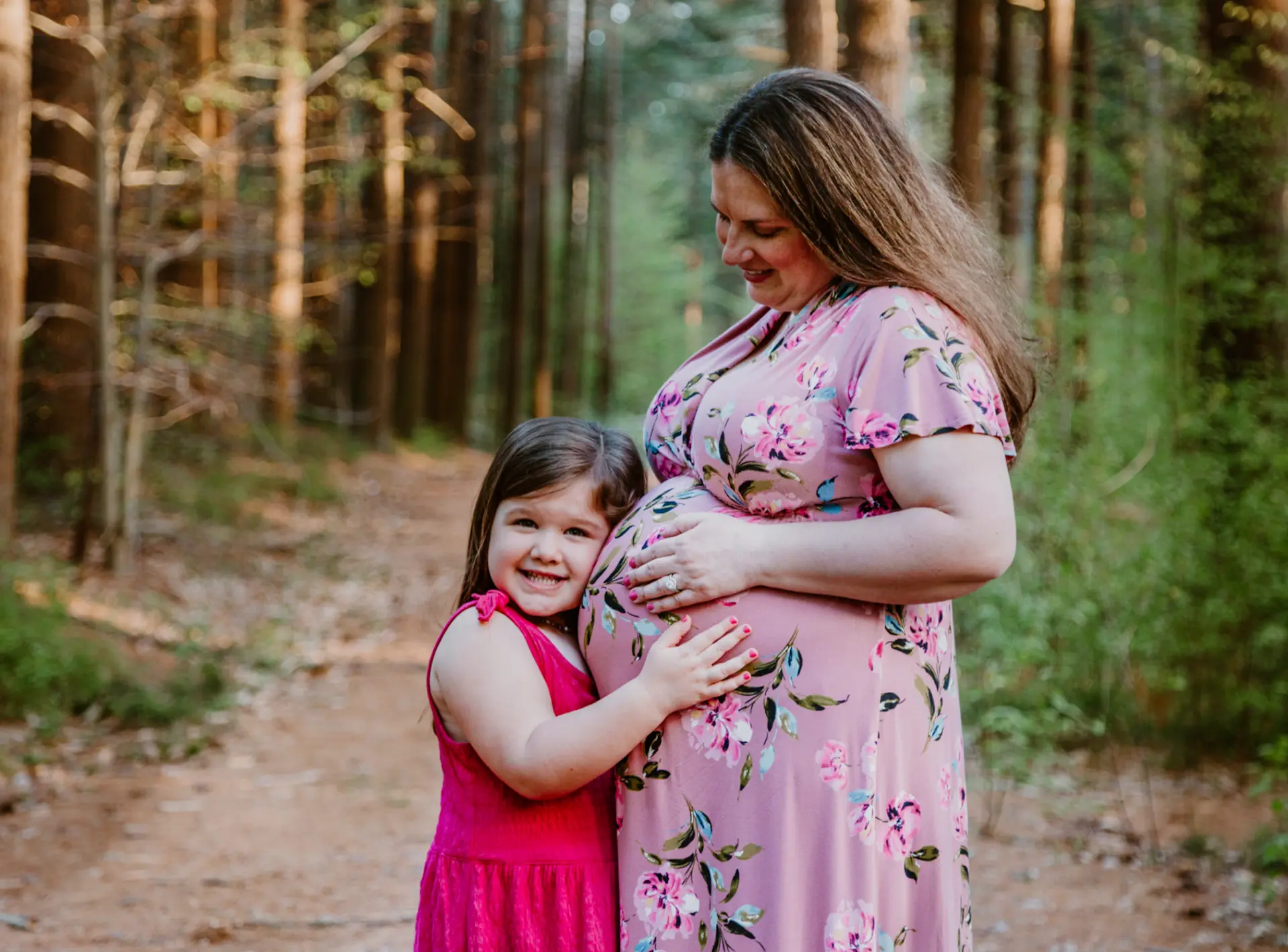 Big sister hugging her mother's baby bump on a pine forest path