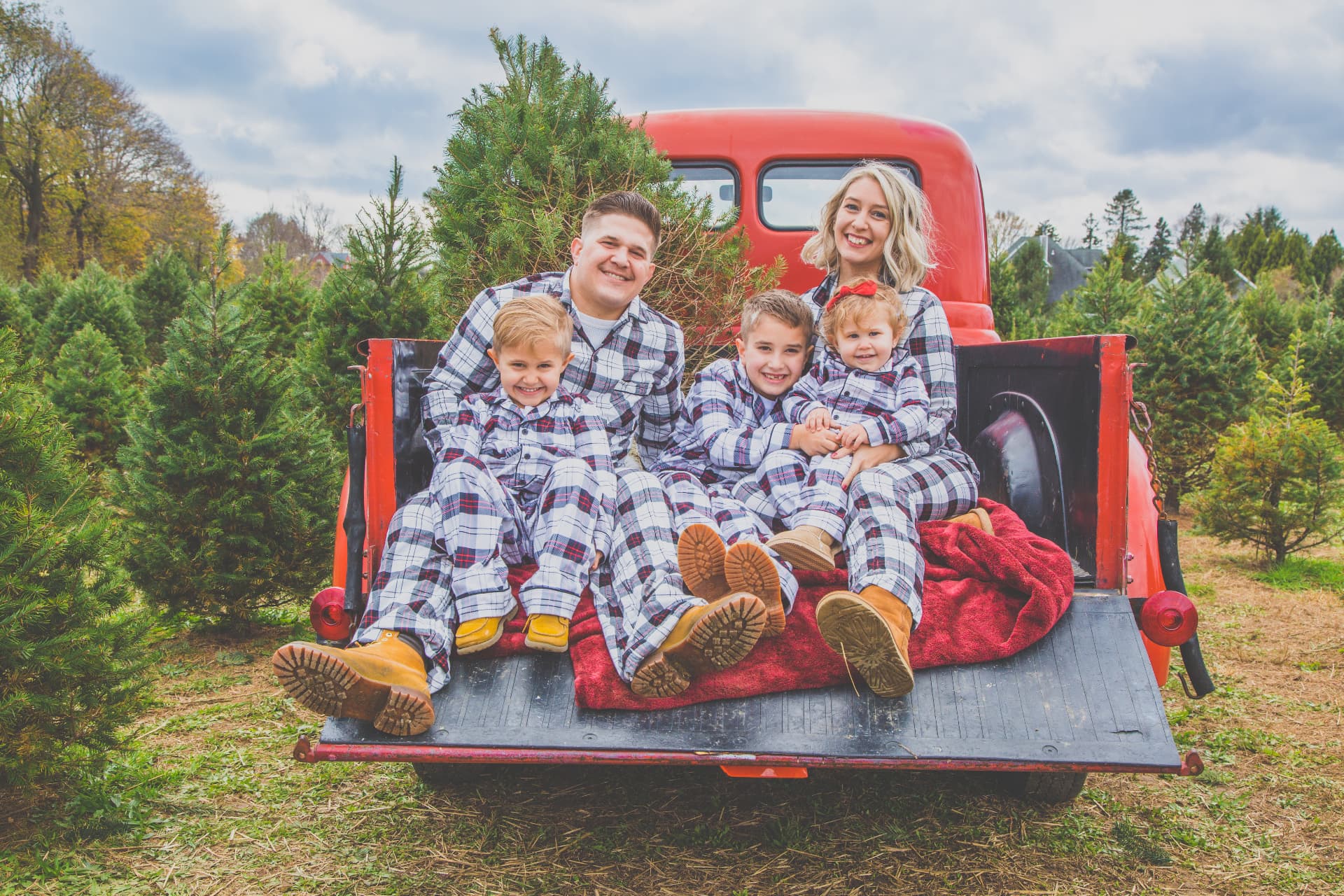 Family of five in matching plaid pajamas in the bed of a vintage red truck at a Christmas tree farm