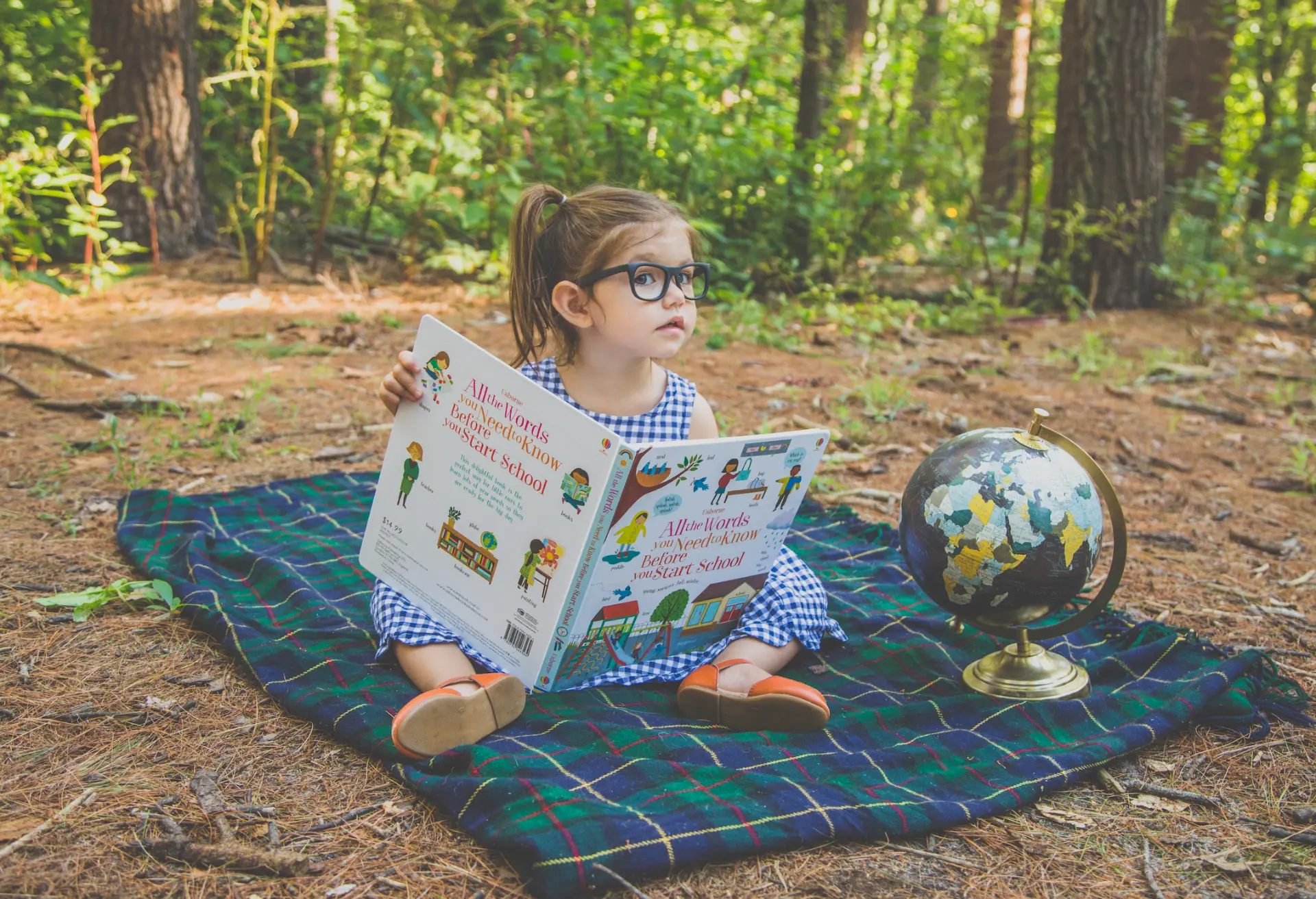 Toddler girl in glasses reading a picture book on a plaid blanket